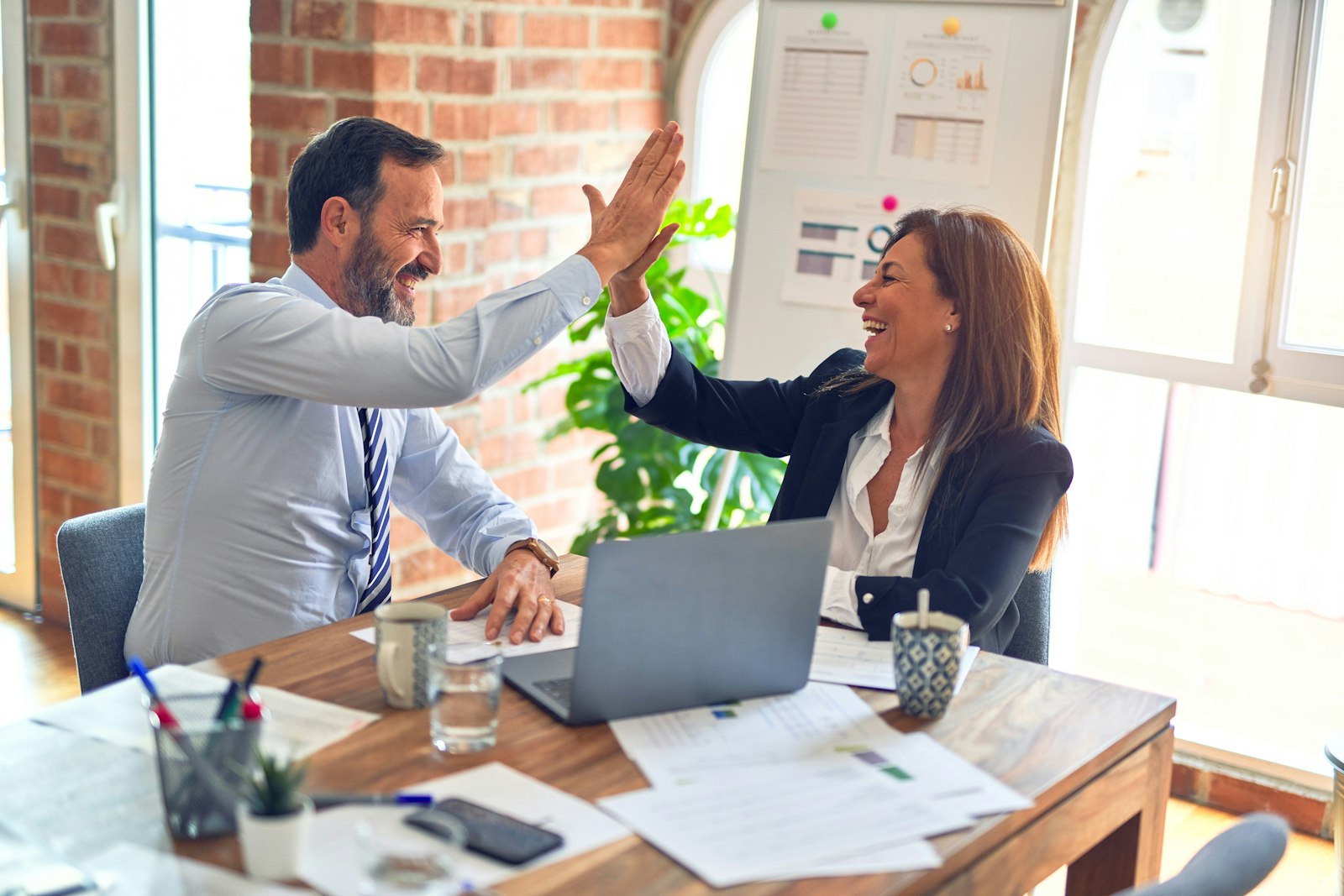 Two business professionals celebrating success with a high five over a laptop and financial reports in a modern office.