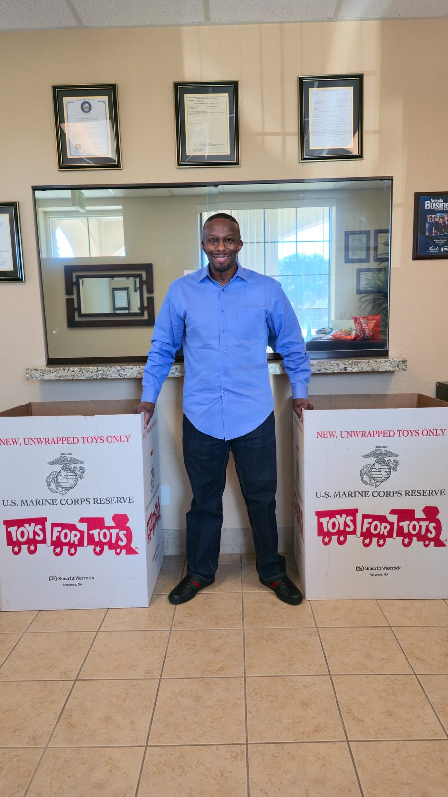 Business Continuity Technologies owner standing between two Toys for Tots donation boxes in the BCT office, supporting the annual community toy drive for children.
